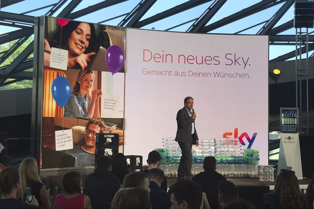 CEO von Sky Deutschland, Carsten Schmidt, präsentiert die CSR-Initiative Sky Ocean Rescue auf einer Bühne in der BMW Welt München. Im Hintergrund eine großflächige Unterwasser-LED-Inszenierung mit Meeresschildkröte und Korallenriff, davor nachhaltige Installationen aus recycelten Plastikflaschen im Rahmen des Event- und Nachhaltigkeitskonzepts.