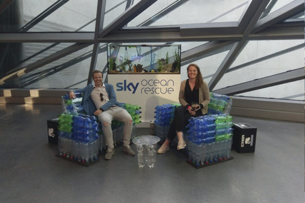 Damian Holmer und Teammitglied auf einer nachhaltigen Installation im Rahmen von Sky Ocean Rescue in der BMW Welt München. Die Sitzmöbel und der Tisch bestehen aus recycelten PET-Flaschen und waren Teil des CSR- und Innovationskonzepts rund um Nachhaltigkeit, Startup-Kultur und Community-Events. Im Hintergrund ist das Branding von Sky Ocean Rescue zu sehen.