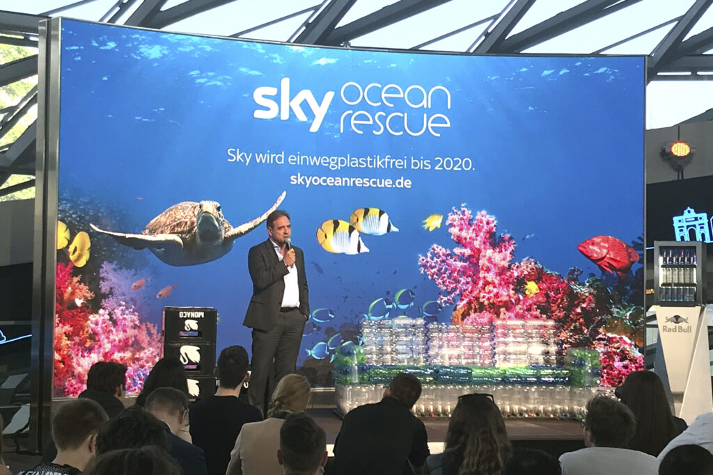CEO von Sky Deutschland, Carsten Schmidt, präsentiert die CSR-Initiative Sky Ocean Rescue auf einer Bühne in der BMW Welt München. Im Hintergrund eine großflächige Unterwasser-LED-Inszenierung mit Meeresschildkröte und Korallenriff, davor nachhaltige Installationen aus recycelten Plastikflaschen im Rahmen des Event- und Nachhaltigkeitskonzepts.
